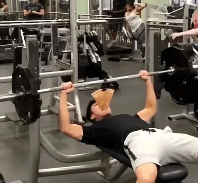 健身房场景中，一名穿黑色T恤和白色短裤的男子正在做卧推训练，双手紧握杠铃的同时，他仰头张嘴，正上方悬浮着一片即将掉落的薯片，似乎在挑战边健身边吃东西的高难度动作。背景中还有其他健身者在使用器械，整个画面充满了幽默和荒诞感。