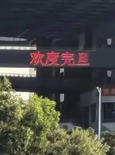 图片中展示了一栋建筑物外墙上的红色LED电子屏，屏幕上醒目地显示着“欢度完且”四个大字，疑似将“元旦”误写为“完且”。背景可见建筑物的结构和前景的绿色树木，整体场景为白天户外。这一错别字可能是由于输入失误或字形相似导致，引发网友对新年祝福标语出错的调侃。