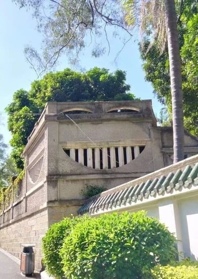 一张展示传统中式风格石墙建筑的照片。画面中央是一座石制结构，顶部有两个圆形窗洞，中间部分有一排竖条形栏杆。建筑右侧连接着一段带有绿色瓦片的传统中式屋檐。周围环绕着茂密的绿色植物，包括高大的树木和修剪整齐的灌木丛。天空晴朗，呈现出明亮的蓝色。石墙底部左侧有一个垃圾桶。整体场景展现了传统中式建筑与自然环境的和谐融合。