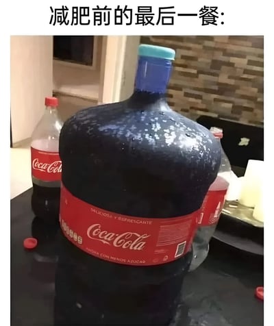 图片上方有文字“减肥前的最后一餐:”，主体画面是一个异常巨大的可口可乐塑料瓶（瓶身有红色标签，印有“Coca-Cola”字样及西班牙语“DELICIOSA Y REFRESCANTE”），瓶身有水珠，周围摆放着普通大小的可口可乐瓶和一些冰块。通过夸张的超大瓶饮料，幽默地表现人们在开始减肥前放纵自己、过度摄入高热量食物的心态，带有调侃和搞笑的意味。