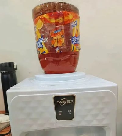 一张展示生活小妙招（或搞笑操作）的照片：一个超大容量装的康师傅冰红茶饮料瓶被直接倒扣在小型浪木牌饮水机顶部，似乎想利用饮水机的结构来倒饮料。瓶身标签清晰可见，饮水机控制面板上有加热和制冷按钮。这种将大瓶装饮料直接放在饮水机上的非常规用法，形成了一种幽默又实用的视觉效果，让人会心一笑。