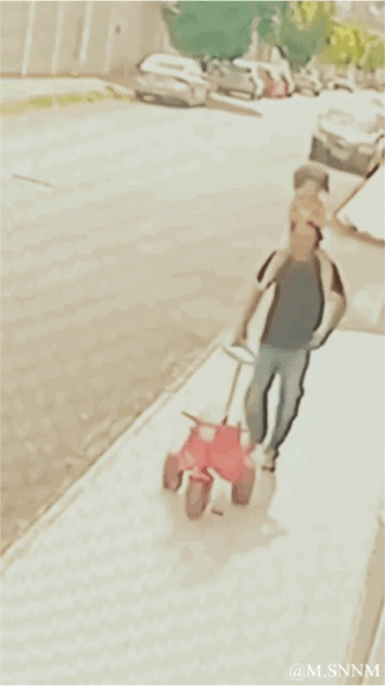 这是一段动态GIF，记录了街头的趣味视觉错位场景。画面中，一位穿着深色上衣、牛仔裤的成年人正推着红色小推车走在人行道上，后方的小孩因为拍摄视角的巧合，看起来像是稳稳坐在成年人的头顶上，制造出荒诞又搞笑的错觉效果，充满生活里的意外趣味。