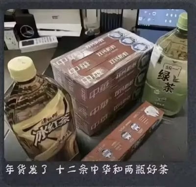 图片展示了一张办公桌场景，桌上摆放着一盒十二条装的中华香烟、一瓶冰红茶和一瓶绿茶。图片底部配有文字“年货发了十二条中华和两瓶好茶”，通过将名贵的中华香烟与常见的瓶装茶饮料（冰红茶、绿茶）并列为“年货”和“好茶”，形成幽默反差，调侃新年福利看似丰厚实则“接地气”的有趣情况。