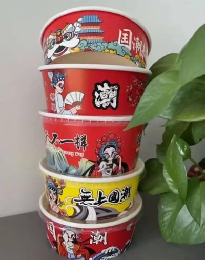 这张图片展示了五个堆叠在一起的国潮风格一次性纸碗，从上到下依次为：红色碗身印有醒狮与古建筑图案，搭配“国潮来”字样；红色碗身印有持折扇的京剧花旦与“潮”字；红色碗身印有戴墨镜的京剧花旦与“天天不一样”字样；黄色碗身印有京剧花旦与“无上国潮”字样；红色碗身印有国风老者与“国潮”字样。纸碗旁有绿色绿植作为衬托，放置在灰色台面上，整体设计融合传统国风元素与潮流风格，色彩鲜明，充满活力。