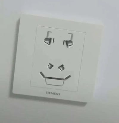 这是一张拍摄于日常生活中的墙壁插座照片，插座为西门子品牌。插座的插孔布局形成了趣味的视觉巧合：上方的两孔插座如同一双眼睛，下方三孔插座的两个斜向插孔像皱起的眉毛，下方的直孔则像咧开的嘴巴，整体组合起来酷似一个带着坏笑的人脸表情，让普通的日常物品变得充满趣味。