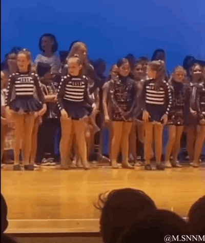 这是一段动态GIF图，呈现了一群穿着统一条纹舞蹈服的小女孩在舞台上准备舞蹈表演的场景。前排的小女孩们起初跟着节奏摆动身体，其中一个小女孩突然停下动作，站在原地露出尴尬的神态，与周围仍在舞动的同伴形成鲜明反差，营造出好笑的社死既视感，让人联想到童年时的尴尬经历。