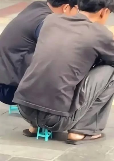 整张图为室外实拍内容，画面中有两名背对镜头的成年男性，均身穿灰调衣物、脚穿拖鞋，以蹲坐的姿势分别坐在一个尺寸极小的浅蓝色迷你塑料凳上，小凳子仅巴掌大小，和成年人的身形形成强烈的反差感，看起来仿佛两人直接蹲在地面上，具有很强的搞笑效果，背景是平整的灰色地砖路面。