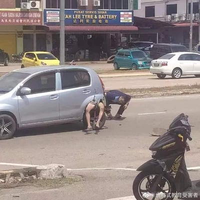这是一张在街边拍摄的静态照片，呈现出极具趣味的视觉错位效果：画面中的银色轿车右后轮旁，两名男子的身体位置恰好重合，其中一人的下半身与另一人的上半身拼接在一起，看起来就像是一个人钻进了车胎里，只露出双腿和臀部，旁边的男子弯腰俯身，似乎在协助修车。背景是马来西亚的街边商铺，包括一家名为“达利轮胎电池”的维修店，还有其他停放的车辆，日常的修车场景因这个巧合的错位变得荒诞又滑稽，让人忍俊不禁。