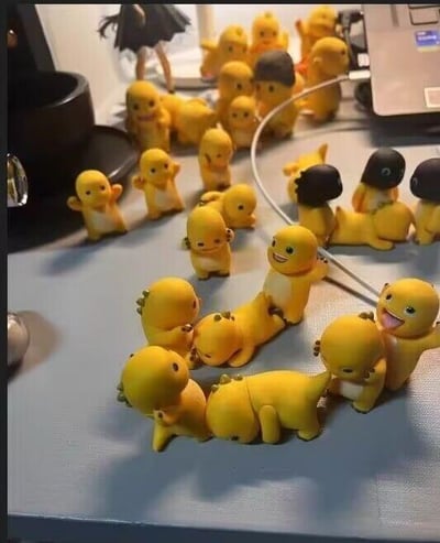 这张实拍图展示了一张桌面上摆满了大量黄色的奶龙手办，这些手办造型各异，有的站立挥手，有的躺卧在地，还有的互相堆叠在一起，形成了热闹又有趣的排列。奶龙是国产动画《奶龙》中的卡通角色，有着圆滚滚的黄色外形和软萌的神态，图中的手办还原了它的各种活泼小表情，有的开心大笑，有的呆萌平静，整体画面充满治愈感，看起来十分可爱。