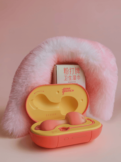 图片展示了一个粉色主题的产品组合，前景是一个黄色和橙色相间的打开的盒子，品牌标识为"PINK PUNCH"，里面装有一个粉色的成人玩具。盒子后面有一个粉色的毛绒物品，形状类似U型或月牙形。背景中可以看到一个白色包装的盒子，上面有红色中文文字"粉打品牌卫生湿巾"。整个画面采用柔和的粉色背景，营造出可爱甜美的视觉效果。