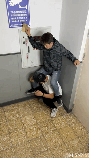 这是一张竖屏实拍的趣味照片，画面中一名穿黑灰迷彩外套的男子，被下方穿白T恤戴黑帽子的男子半蹲托举着，借助对方的高度去够墙上高处的电箱柜门。由于视觉错位，看起来像是迷彩外套男子骑在对方脖子上，制造出搞笑的视觉效果。画面左上角还有一块蓝色的“24小时视频监控”标识牌，提示此处处于监控区域。