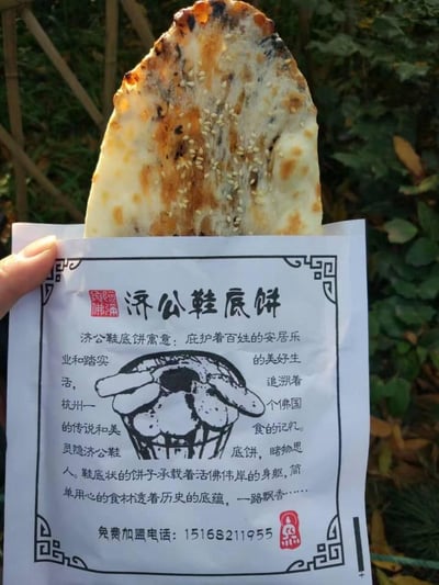 这张图片展示了杭州灵隐的特色街头小吃济公鞋底饼，饼身呈类似鞋底的长形，表面撒有白芝麻，经过烘烤后带有自然的焦色，看起来香气十足。饼被放置在印有品牌宣传内容的白色带边框包装袋中，背景是户外的绿色植物。包装袋上印有济公形象的简笔画，搭配详细的文字介绍，将这款小吃与杭州灵隐的济公传说相结合，赋予其庇护百姓安居乐业的美好寓意，称其承载着佛国传说与美食记忆，用简单食材传递历史底蕴，同时标注了免费加盟电话，兼具美食展示与商业推广的属性。