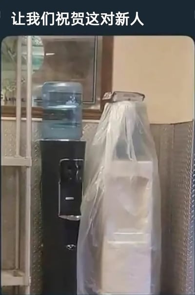 图片展示了两台并排放置的饮水机，左侧是黑色机身配蓝色水桶，右侧白色饮水机被透明塑料袋覆盖，袋口自然垂落形似婚纱裙摆。图片上方配有文字'让我们祝贺这对新人'，通过拟人化手法将日常办公用品幽默地塑造成新婚夫妇形象，营造出荒诞又喜庆的喜剧效果。