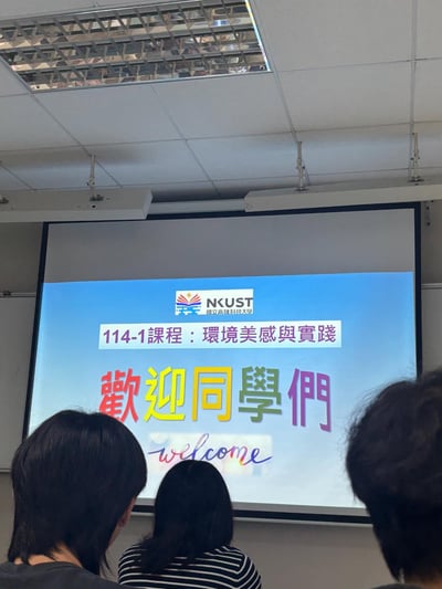 教室场景，三名学生背影面向前方投影屏幕。屏幕上显示高雄科技大学(NKUST)的114-1课程「环境美感与实践」欢迎页面，主要文字为彩色的「歡迎同學們」及英文「welcome」。教室天花板有格栅式灯具和投影仪设备。