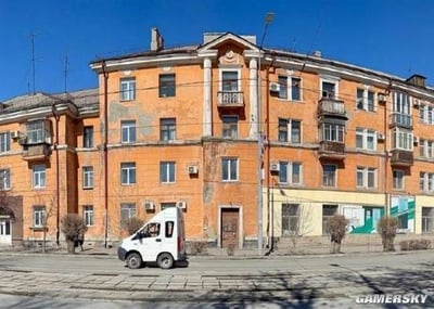 图片展示了一栋橙色的老式多层公寓楼，建筑外墙存在部分油漆剥落的痕迹，带有古典风格的立柱和装饰性山花。楼体分布着多个窗户和阳台，部分阳台安装了空调外机。楼前街道上停放着一辆白色面包车，周围有几棵无叶树木，地面为石板铺就的路面。背景是晴朗的蓝色天空，画面右下角可见“GAMERSKY”水印标识。整体呈现出写实的城市街景氛围，建筑风格具有年代感。
