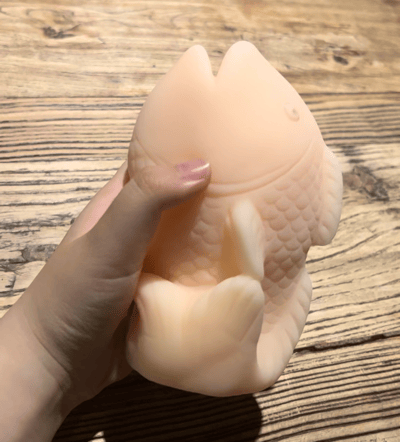 一只手正握着一个米白色的鱼形解压玩具，玩具表面有细腻的鱼鳞纹理，整体呈现出柔软的质感。背景是带有自然木纹的桌面，光线柔和，突出了玩具的立体感和材质特性。这是一款典型的捏捏乐减压玩具，设计成可爱的鱼类造型，适合日常解压使用。
