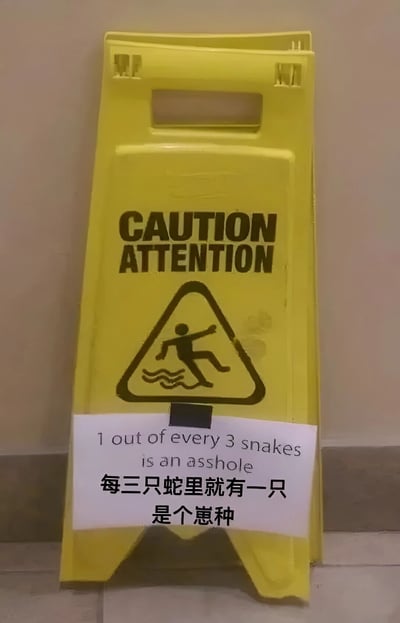 一张黄色的标准警示牌，原本用于提示“小心地滑”（牌面有英文“CAUTION ATTENTION”和一个人滑倒的图案），但被人在下方贴了一张白色贴纸。贴纸上用英文写着“1 out of every 3 snakes is an asshole”，下方配有中文翻译“每三只蛇里就有一只是个崽种”，通过一本正经的警示形式搭配无厘头的蛇类调侃，形成强烈反差萌的搞笑效果。