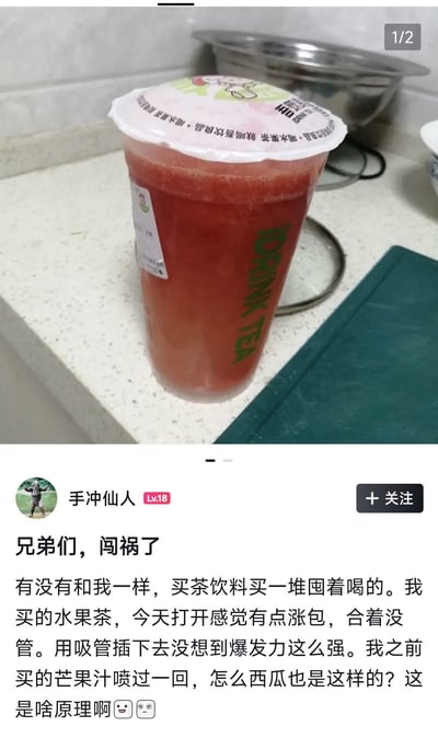 这是一张社交媒体截图，整体分为上下两部分，上半部分实拍了一杯放置在厨房石英石台面上的超大杯西瓜水果茶，杯子是透明塑料杯，杯身印有绿色的“DRINK TEA”字样，杯盖印有品牌标语，杯内的红色果茶因为产气已经明显膨胀涨袋，杯盖被顶得凸起；下半部分是等级Lv.18的用户“手冲仙人”发布的帖子文字，用户称自己有囤购茶饮慢慢喝的习惯，这次囤的水果茶打开前就发现涨包，没在意直接插吸管，结果果茶因为内部压力大直接喷溅出来，他之前囤的芒果汁也发生过同样的情况，因此好奇提问这是什么原理，帖子末尾附带两个表情，右上角标注该图是组图的第一张（1/2）。