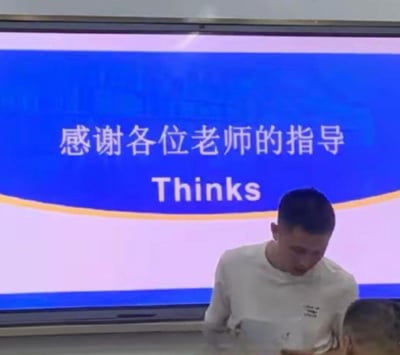 这是一个答辩或汇报现场的实拍画面，背景的PPT感谢页上，中文写着“感谢各位老师的指导”，但对应的英文却出现拼写错误，将正确的“Thanks”误写为“Thinks”。画面下方的汇报者穿着白色上衣，正低头，似乎并未察觉到这个失误，社死感拉满，是很典型的校园/职场汇报小失误社死场景。