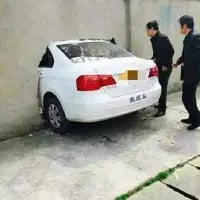这是一张利用拍摄角度制造出视觉错位效果的趣味照片，画面中的白色大众朗逸轿车，从拍摄视角看仿佛整个车头都钻进了前方的墙体中，只露出完整的车尾部分。两位身着黑衣的男子站在车旁，正低头看向车辆，脸上带着诧异、无奈的神情，让这个错位场景显得更加荒诞搞笑，乍一看像是发生了离谱的交通事故，实则是角度带来的错觉。