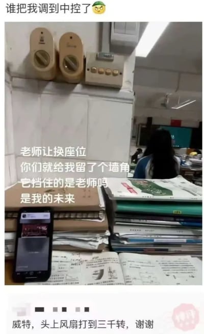 这是一张来自学生视角的教室实拍图，拍摄者的座位被安排在紧邻墙面的角落位置，墙面装有两个风扇旋钮开关和一个控制装置，桌上堆满了中学阶段的课本、练习册与一部亮着屏幕的手机，前方的墙面完全挡住了拍摄者的部分视线。图中添加了多段吐槽文字：顶部用“谁把我调到中控了😤”的玩笑话，把墙角座位调侃成类似“中控”的特殊位置；中间文字“老师让换座位，你们就给我留了个墙角，它挡住的是老师吗，是我的未来”，用夸张的语气吐槽这个座位的糟糕，将视线被挡的情况上升到“挡住未来”的搞笑程度；底部还有一条类似评论的文字“威特，头上风扇打到三千转，谢谢”，进一步用夸张的要求来表达对这个座位的不满。整体内容充满学生时代的趣味吐槽感，用幽默的方式抒发被分到不理想座位的无奈情绪。