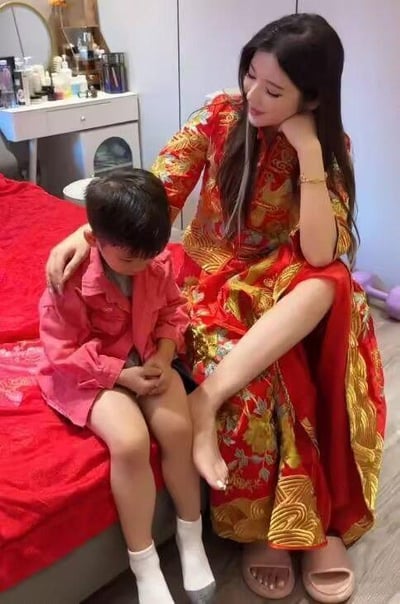 这是一张在卧室拍摄的生活照片，画面中一位身着红底金纹传统服饰的女子坐在床边，她手肘撑在膝盖上，手托着脸颊，另一只手轻轻搭在身旁小男孩的肩膀上，目光温柔地看向小男孩。小男孩穿着粉色上衣，坐在床边，低着头，双手放在腿间，神情显得有些低落。背景里可以看到摆放着各类化妆品的白色梳妆台，床铺铺着红色床单，整体是温馨的居家环境，但小男孩的低落状态让画面多了一丝让人在意的氛围。