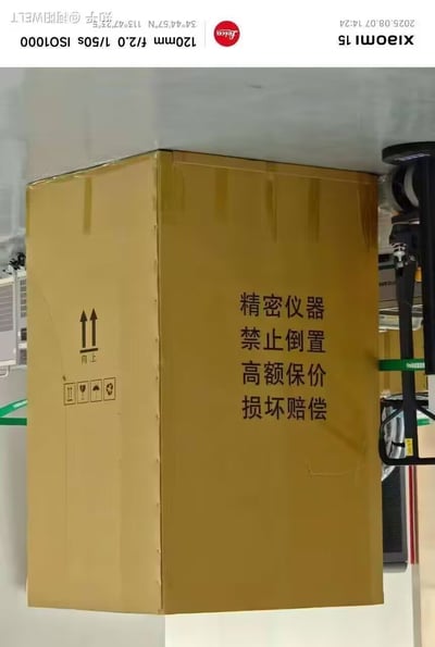 图片展示了一个棕色纸箱，箱体正面印有黑色文字“精密仪器 禁止倒置 高额保价 损坏赔偿”，左侧面有向上箭头标识（标注“向上”）及多个警示图标（含小心轻放、怕水、怕晒、怕压符号）。箱子正被机械吊装设备吊起，处于室内仓储或物流转运场景中，整体画面清晰呈现了贵重精密仪器的运输包装规范要求。