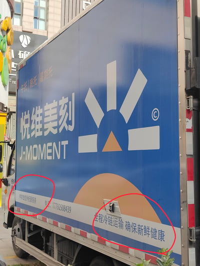 这是一张在城市街道拍摄的厢式货车车身广告图，货厢主体为深蓝色，印有白色的'悦维美刻 J-MOMENT'品牌标识，上方有浅棕色的'午托|晚托|暑期托'字样，表明该品牌同时提供学生托管服务。货厢底部的白色条带区域，标注了'中学生校外托管服务'、联系电话'17792508439'，以及'全程冷链运输 确保新鲜健康'的冷链运输服务宣传语，显示该品牌兼具托管与冷链配送的业务。背景是普通的城市商业建筑，货车为品牌自有配送车辆，用于线下流动宣传。