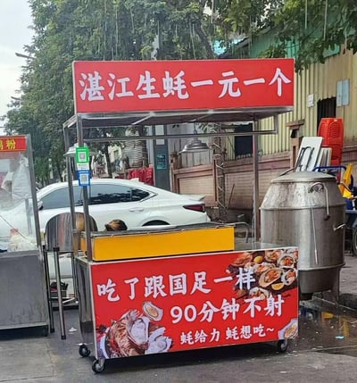 这是一张街头实拍的湛江生蚝小吃推车照片，推车上方红色招牌写着「湛江生蚝一元一个」，下方的红色宣传牌则用玩梗的文案吸睛：「吃了跟国足一样90分钟不射」，还有「蚝给力 蚝想吃~」的谐音标语。

**梗说明**：
1. 双关梗：「90分钟不射」一方面对应足球常规比赛时长，调侃国足在赛事中的表现，另一方面暗喻生蚝的滋补功效；
2. 谐音梗：「蚝给力」「蚝想吃」是「好给力」「好想吃」的谐音，巧妙结合生蚝的「蚝」字，强化产品关联。