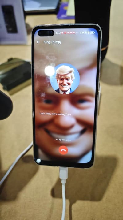 图片展示了一部正在充电的智能手机，屏幕上显示着一个模拟通话界面。联系人名为"King Trumpy"，配有特朗普的头像。通话界面显示"AI speaking..."状态，并有文字"Look, folks, we're making Trum"。这似乎是一个使用AI语音合成技术模拟特朗普声音的应用程序界面，带有幽默和调侃的意味。