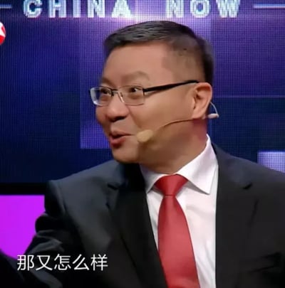 这是一张出自《这就是中国》节目的视频截图，画面中的人物是学者张维为，他身着深色西装、白衬衫与红色领带，佩戴着耳麦，侧头看向画面一侧，嘴巴微张，面部神情略带不屑，仿佛正在强硬回应。画面下方配有白色字幕‘那又怎么样’，背景屏幕上显示着英文‘CHINA NOW’。这张图被网友截取后，成为用来表达无所谓、不在意，或是面对质疑时强硬回应情绪的表情包素材。