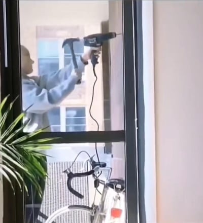 这是一张利用窗户玻璃形成趣味视觉错位的照片：窗外的人一只手握着电钻、另一只手拿着锤子，透过室内的玻璃窗看过去，电钻的钻头恰好对准室内的墙面，营造出「工人正在钻室内墙壁」的错觉，画面充满乌龙感，让人第一眼容易看错，是生活里巧合造就的搞笑趣味场景。
