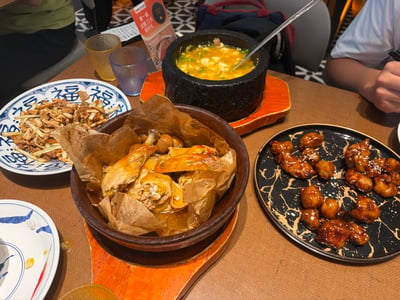 这是一张餐厅聚餐场景的实拍照片，餐桌为棕色桌面，摆放了多道丰盛的菜肴：正中靠前位置是陶制大碗盛装的纸包烤鸡，鸡肉色泽油亮；烤鸡后方是黑色石锅盛装的黄色浓汤羹，表面点缀有绿色葱花，锅内放有金属汤勺；左侧是印有蓝色福字纹样的白瓷盘，盛放着葱丝搭配的炒制肉菜；右侧是带有金色裂纹纹理的黑色餐盘，盛放着裹有亮泽糖醋酱汁的炸制菜肴。桌面还摆放有磨砂质感的玻璃杯、餐厅活动提示立牌，桌边露出两名就餐者的部分身体，其中一人手持筷子，整体氛围轻松，是朋友共同享用美食的场景。
