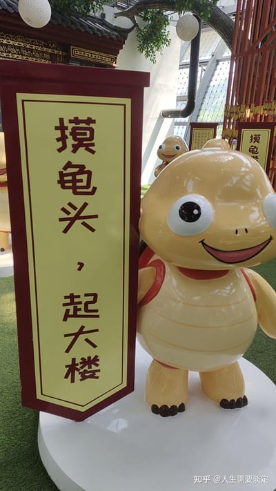 图片中展示了一个黄色卡通乌龟雕像，它有着大大的圆眼睛、微笑的嘴巴和红色的龟壳边缘，正站立在白色底座上。乌龟旁边立着一个红底黄面的竖幅标语，上面写着“摸龟头，起大楼”。背景中可以看到传统风格的建筑装饰、绿色植物以及其他类似的标语牌，整体场景像是在一个室内文化展览或主题场馆中。图片底部有水印“知乎@人生需要淡定”。