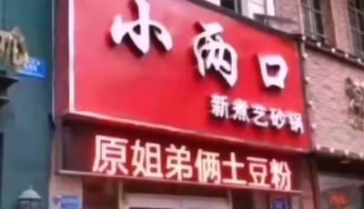 图片展示了一家店铺的红色招牌特写，招牌主体为红色底色，上方有白色大字“小两口”，下方标注“新煮艺砂锅”，底部另有一块白色文字招牌写着“原姐弟俩土豆粉”。背景可见部分建筑物的砖墙和窗户，整体为真实拍摄的临街店铺门面场景。