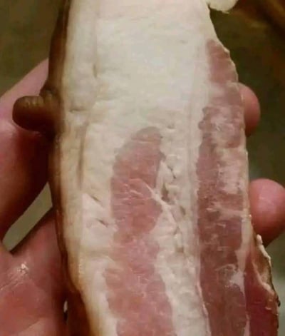 这是一张利用视觉错位效果的实拍图片，主体是一块肥瘦分层清晰的五花肉（看起来像是经过腌制的腊肉类食材），其左侧边缘的凸起结构和整体被握持的视角，让观察者第一眼容易误以为是一只手握住了这块肉，但实际上这个类似“手指”的部分是肉本身的边缘形态，形成了食材和人手混淆的趣味视觉错觉，极具迷惑性和趣味性，能让人感受到日常事物中的意外趣味。