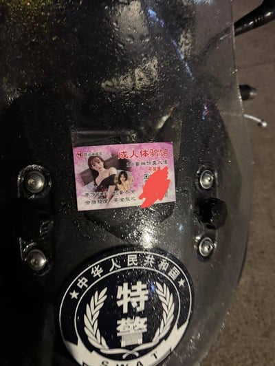 这张图拍摄的是带有「中华人民共和国 特警」标识的黑色装备（疑似防护盾或头盔），其表面被违规张贴了一张成人体验馆的低俗小广告。小广告标注「18+禁止未成年」，主打「1:1高端仿真人偶」，附带女性模特图片、宣传语「干净卫生 配套齐全 合法经营 安全放心」以及被涂抹的联系方式。下方的官方特警标识与上方的低俗小广告形成强烈的荒诞反差，凸显了这类「牛皮癣」广告的无孔不入。