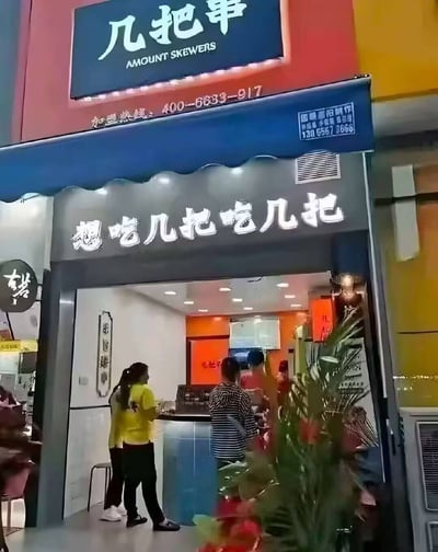这是一家烧烤店“几把串（Amount Skewers）”的门面照片。店铺名称和门头宣传语“想吃几把吃几把”利用了中文的谐音梗，带有明显的幽默和恶搞气质，同时配合直白的英文翻译，令人会心一笑。画面中有顾客进店，门口摆有开业花篮，整体氛围轻松，有趣，极具网络传播特色。这种谐音和口语表达会让店铺名字广为流传，适合喜欢网络梗与恶搞文化的年轻群体。