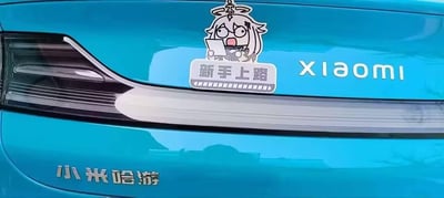 这是一张青蓝色小米汽车尾部的实拍图，车主对车辆进行了个性化的整活改装：保留了车尾的“Xiaomi”英文标识，将下方的品牌字样改为“小米哈游”，玩起了小米与米哈游的联动梗；同时在车尾中部贴有结合《原神》角色派蒙的“新手上路”车贴，派蒙呈现出惊讶慌张的表情，手持纸张，搭配警示文字，既起到提醒后车的作用，又融入了二次元游戏元素，整体内容充满趣味整活的氛围，展现了车主的二次元喜好与创意改装的巧思。
