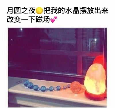 图片上方配有文字'月圆之夜🌕把我的水晶摆放出来改变一下磁场💖'，下方是一张实拍照片，展示了窗台场景：摆放着一串蓝色珠子、一个橙色球状物体和一个发光的黄色块状物体（疑似盐灯）。整体通过幽默的方式调侃了在月圆之夜摆放水晶试图改变磁场的行为，带有轻松诙谐的生活气息。