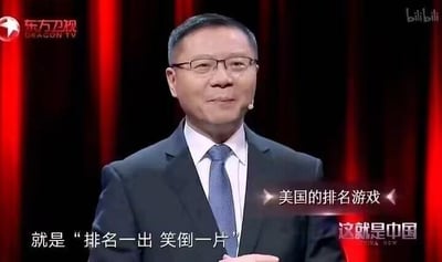这是东方卫视政论节目《这就是中国》的视频截图，画面中的人物是学者张维为，他身着西装、戴着眼镜，面带微笑站在舞台上，似乎正在进行演讲。画面配有字幕“美国的排名游戏”以及“就是‘排名一出 笑倒一片’”，以诙谐的语气调侃美国相关的排名，整体构图清晰，信息明确。