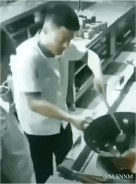 这是一段动态GIF，画面呈现的是餐厅后厨场景，一位身着浅色上衣的厨师正手持锅铲和炒锅进行烹饪。由于视觉错位的效果，锅铲的位置看起来恰好对准厨师的头顶，形成了仿佛厨师在用锅铲铲自己脑袋的搞笑错觉，画面充满滑稽感，让人忍俊不禁。