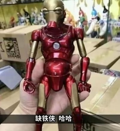 整张图片为实拍场景，一只手握着一款山寨魔改的钢铁侠手办，该手办虽采用了钢铁侠标志性的红金配色和胸口反应炉设计，但整体造型严重走形，头部设计怪异突兀，质感粗糙劣质，和正版钢铁侠形象相差甚远。图片底部配有黑底白字的配文“缺铁侠 哈哈”，利用“钢铁侠”的谐音玩梗，调侃这款劣质山寨手办缺失了钢铁侠的核心设计精髓。背景可见堆放的纸箱和其他陈列的手办，推测拍摄场景为玩具售卖店铺或者手办仓库。