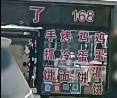 这是一张国内街头小吃店的玻璃门实拍图，门面上用红色字体分四列竖排展示了四种餐品：手抓饼、烤冷面、鸡蛋饼、鸡蛋灌饼，但横排阅读时会看到“手烤鸡鸡”“抓冷蛋蛋”等搞笑的组合，形成了无心的视觉趣味效果。玻璃门上方还有一个单独的“了”字和数字“168”，门的下方贴有多个收款二维码，整体是非常接地气的街边小店风貌，没有谐音或双关梗，只是文字排列方式造成的趣味错觉。