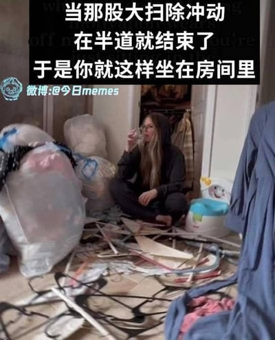 图中展示一位长发女性穿着深色连帽衫，盘腿坐在凌乱的房间地板上，手中拿着玻璃杯正在喝水。房间内散落着大量衣架、衣物、大型毛绒玩具和一个带卡通图案的小垃圾桶，整体环境显得杂乱无章。上方文字“当那股大扫除冲动在半道就结束了 于是你就这样坐在房间里”幽默地描绘了人们常有的经历：兴致勃勃开始大扫除，却在中途失去动力，最终只能无奈地坐在更乱的房间里休息。
