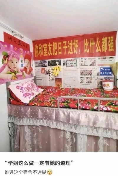 这张图片展示了大学宿舍的一个上铺区域，布置极具复古年代感：铺着鲜艳的大红花图案床单，床头墙面悬挂着写有“你和室友把日子过好，比什么都强”的红色励志横幅，墙面上还贴着老式的“研究生宝宝”海报、报纸，甚至挂着一串香蕉，右侧还有“禁止大小便”的标识，整体风格仿佛复刻了上世纪的家居场景。图片下方配有调侃文字：“学姐这么做一定有她的道理”“谁进这个宿舍不迷糊😳”，用反差感的幽默表达了对这种独特宿舍布置的惊讶与好笑，引发网友对宿舍生活趣味场景的共鸣。