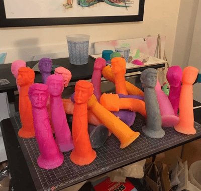 图片展示了一个工作室场景，桌子上摆放着多个色彩鲜艳的雕塑作品。这些雕塑呈现出柱状形态，顶部雕刻有人脸造型，颜色包括粉色、橙色、紫色、灰色等多种鲜艳色调。雕塑被随意地堆放在带有网格图案的切割垫上，背景中可以看到一些制作材料和工具，包括蓝色花纹的杯子、颜料和其他未完成的作品。墙上还挂着一幅装饰画，整个场景看起来像是一个手工艺人的工作空间。