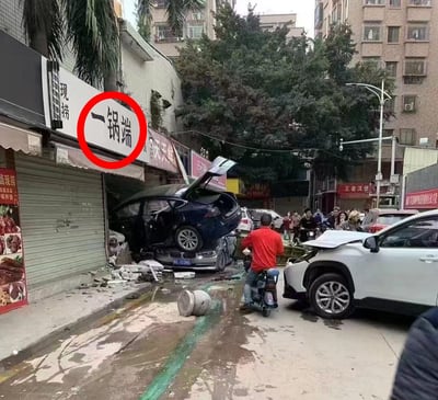 照片展示了一起交通事故现场：一辆黑色轿车撞破街边店铺卷帘门冲入店内，车头部分嵌入建筑，车尾翘起；其下方还叠压着另一辆银色轿车，两车均严重受损。店铺上方招牌写有“一锅端”字样，被红色圆圈重点标注。周围街道聚集多名围观群众，背景可见居民楼、绿树及“王者汉堡”等商铺招牌，现场散落碎石与车辆碎片。
此处存在文字与场景的巧合幽默：“一锅端”原指一次性全部处理或抓捕，图片中车辆堆叠碰撞的混乱场面恰似“一锅端”的字面视觉化呈现。
