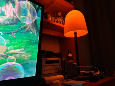 这是一张室内私人桌面的实拍图，左侧显示器正在播放带有梦幻泡泡元素的日系动画场景，画面清新治愈；右侧暖黄色台灯散发柔和光线，照亮了桌面的雅马哈音响、小瓶饮品、遥控器等物品，上方木质玻璃柜内陈列着手办与容器，整体氛围温暖闲适，展现了适合放松的深夜个人休闲空间。