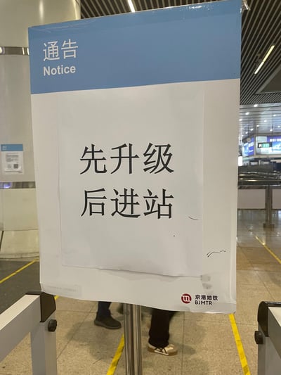 这是一张在京港地铁站内拍摄的官方通告牌照片，蓝色顶部区域标注“通告 Notice”，白色主体区域用醒目大字提示“先升级 后进站”，右下角带有京港地铁（BJMTR）的标识。通告牌设置在地铁站闸机附近，地面可见黄色引导线，背景能看到站内的设施与环境，用于提醒乘客需完成相关升级操作后方可进站乘车。